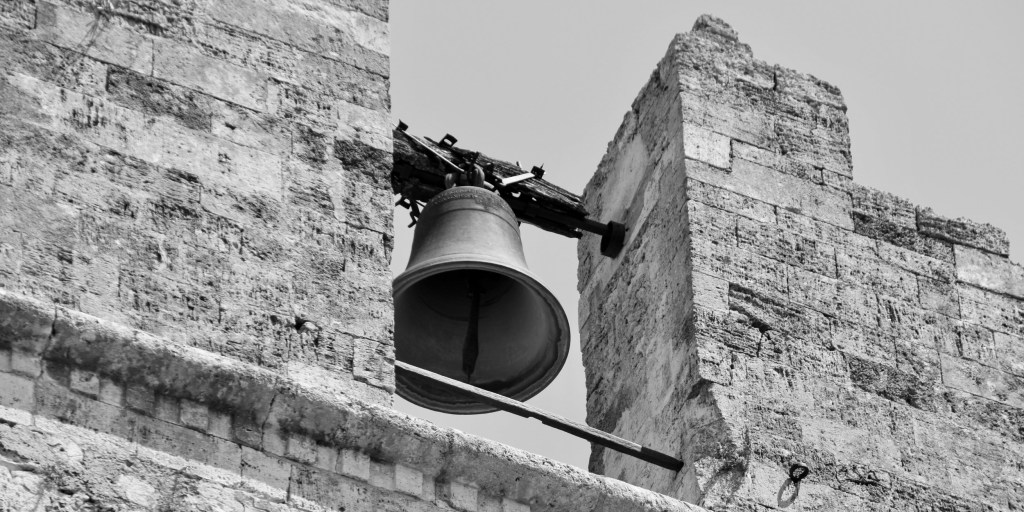 A large bell hangs between two weathered stone structures against the sky.