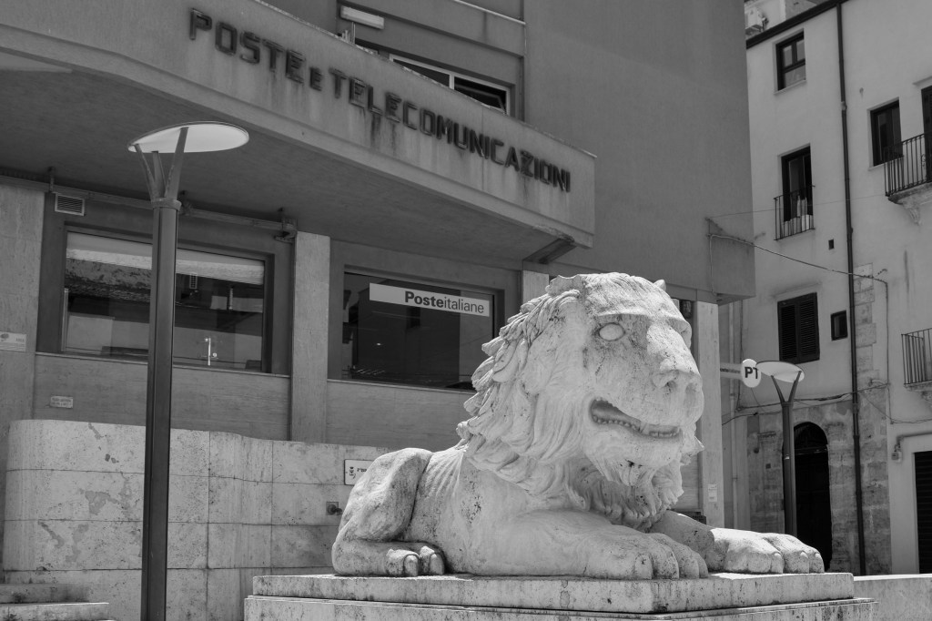 A weathered stone lion lies on a pedestal outside the modern Alcamo post office.