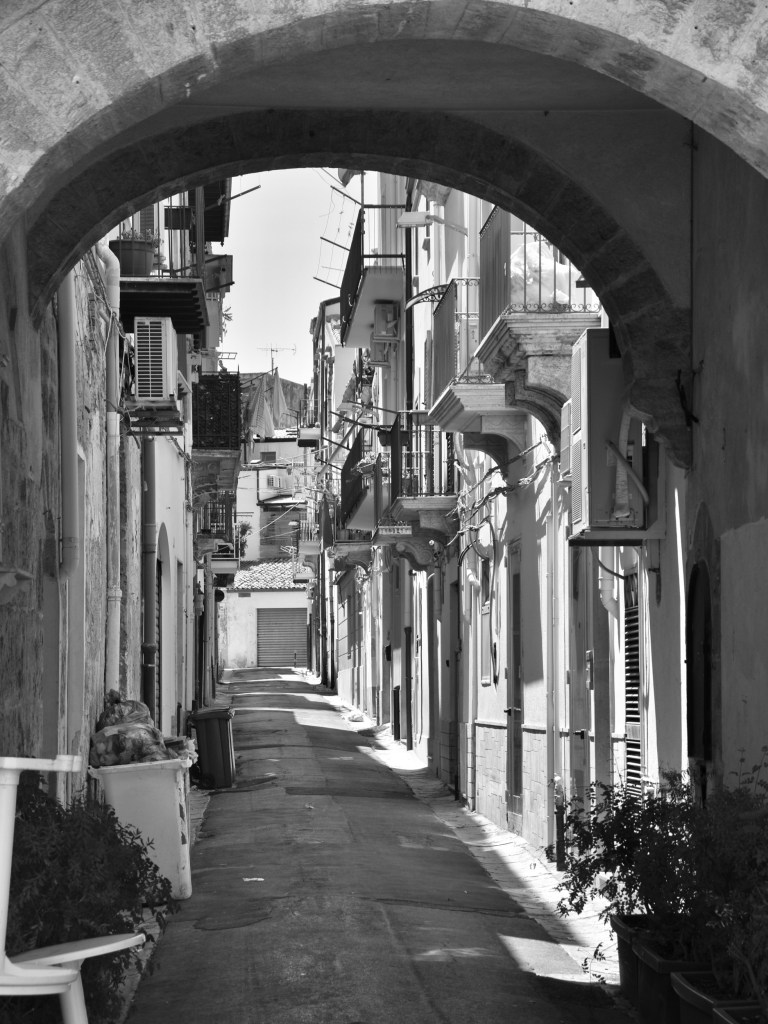 A broad stone arch frames a narrow residential street lined with balconies, air-conditioning units and potted plants.