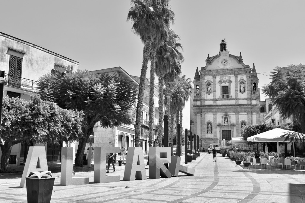 Large freestanding letters spelling “Alcart” stand in a palm-lined square facing an ornate church façade.