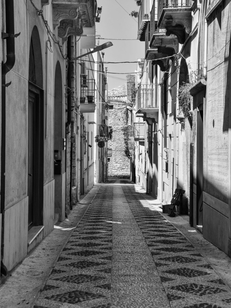A narrow sloping lane with patterned paving runs between closely spaced houses, balconies and overhead cables.