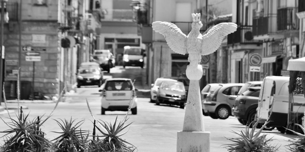 A white winged sculpture rises among agave plants before a street filled with cars.