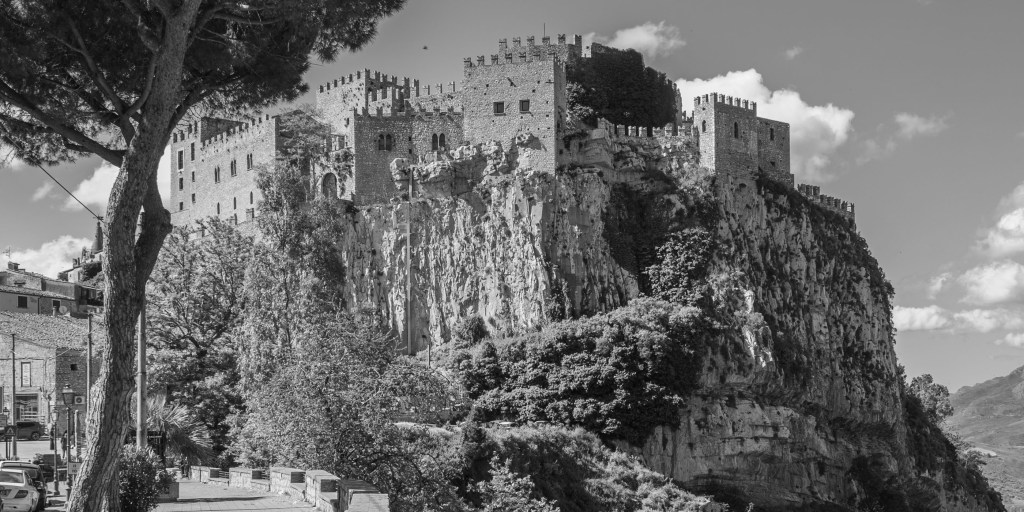 Caccamo Castle rises above a sheer rocky outcrop, framed by a large pine tree and distant mountains.