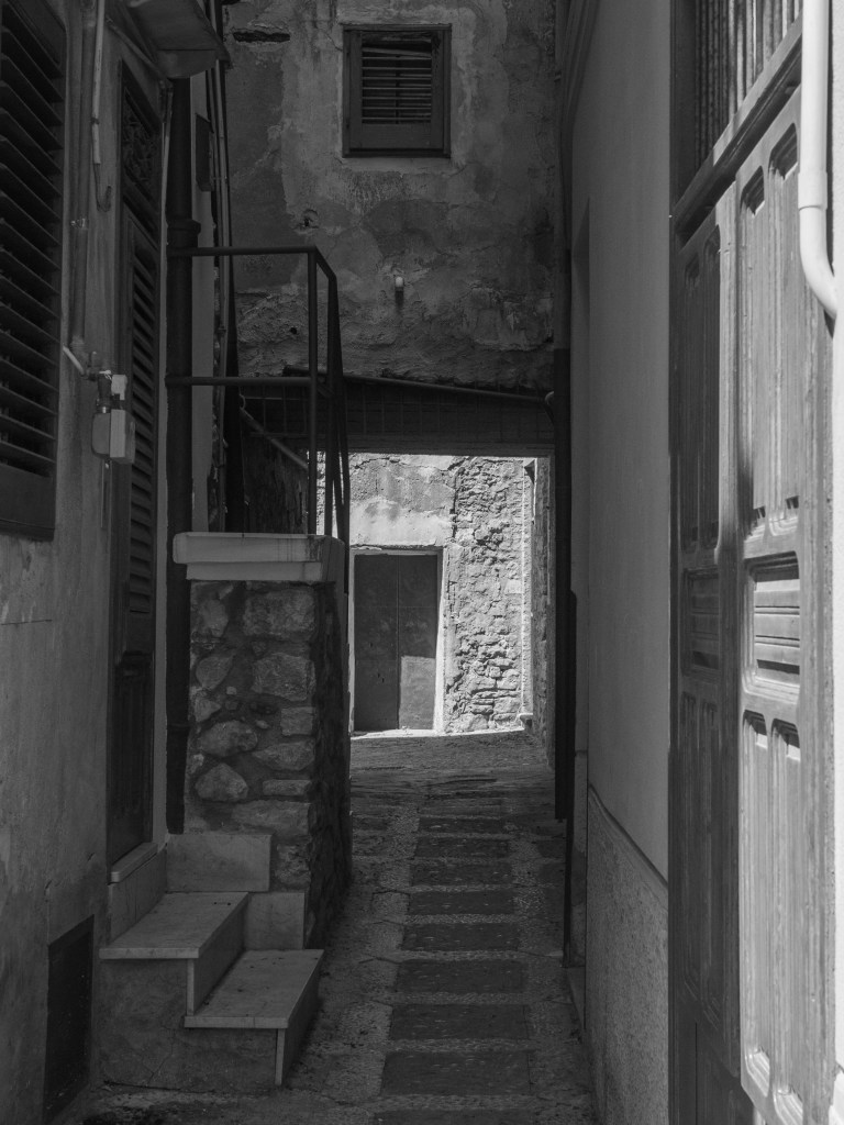 A narrow shadowed passage between buildings leads towards a sunlit stone doorway at the far end.