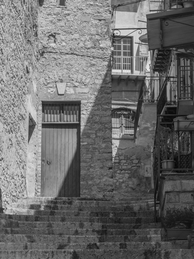 Stone steps rise towards a tall wooden door in a rough masonry wall, with balconies crowded along the right side.