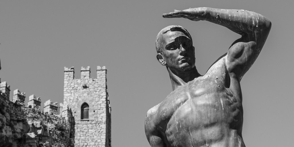 A bronze male figure raises one arm above his face, with the crenellated towers of Caccamo Castle behind him.