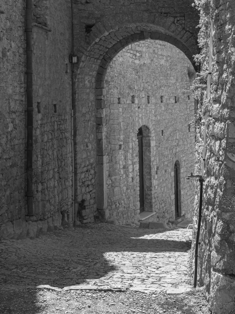 A cobbled passage curves beneath a broad stone arch, with sunlight falling across the ground and walls.