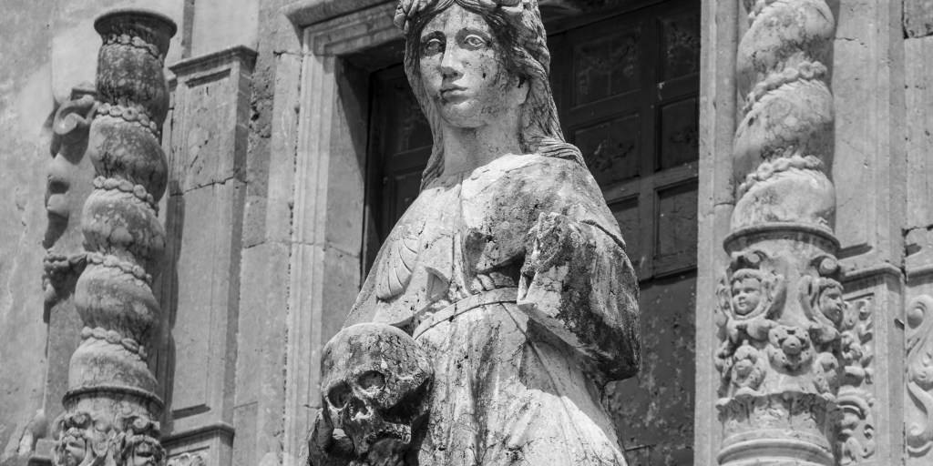 A weathered stone female figure rests one hand on a skull before an entrance flanked by elaborately carved columns.