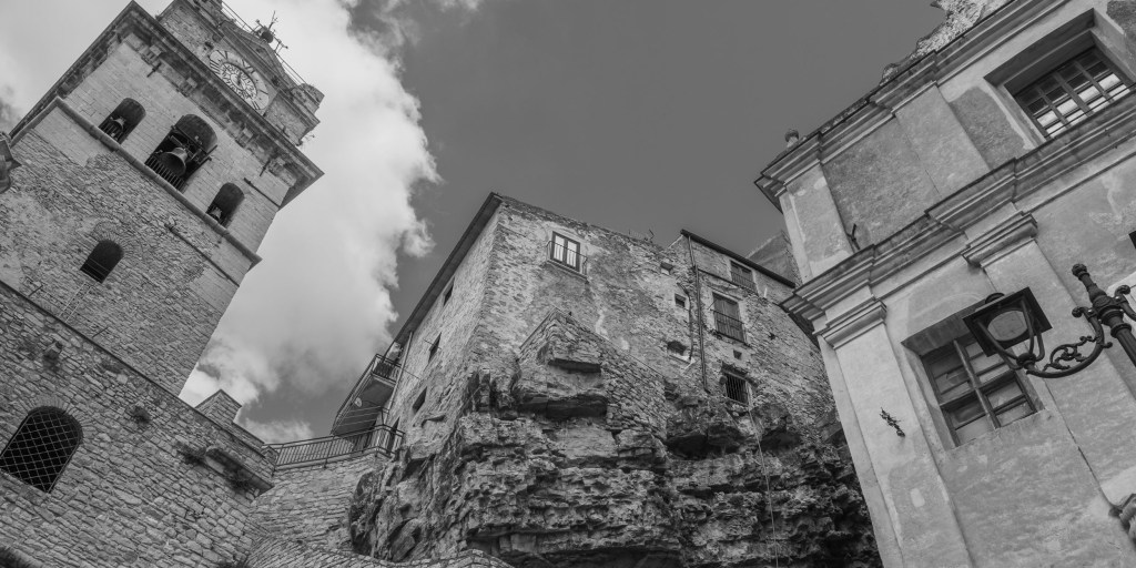 A low-angle view looks up between a clock tower, buildings constructed above exposed rock and a church façade.