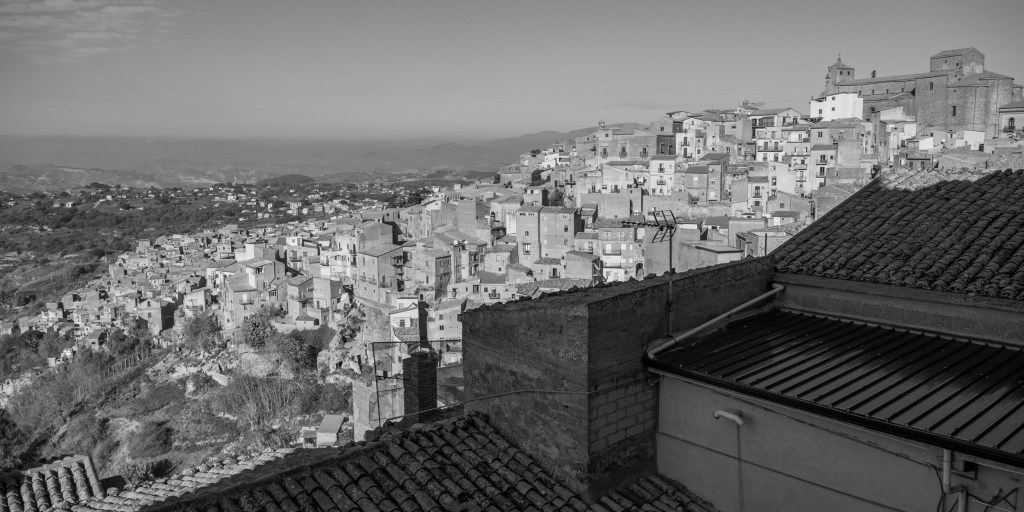 A panoramic view of Calascibetta spreading across a steep ridge, with clustered houses, tiled roofs and a large church above the town.