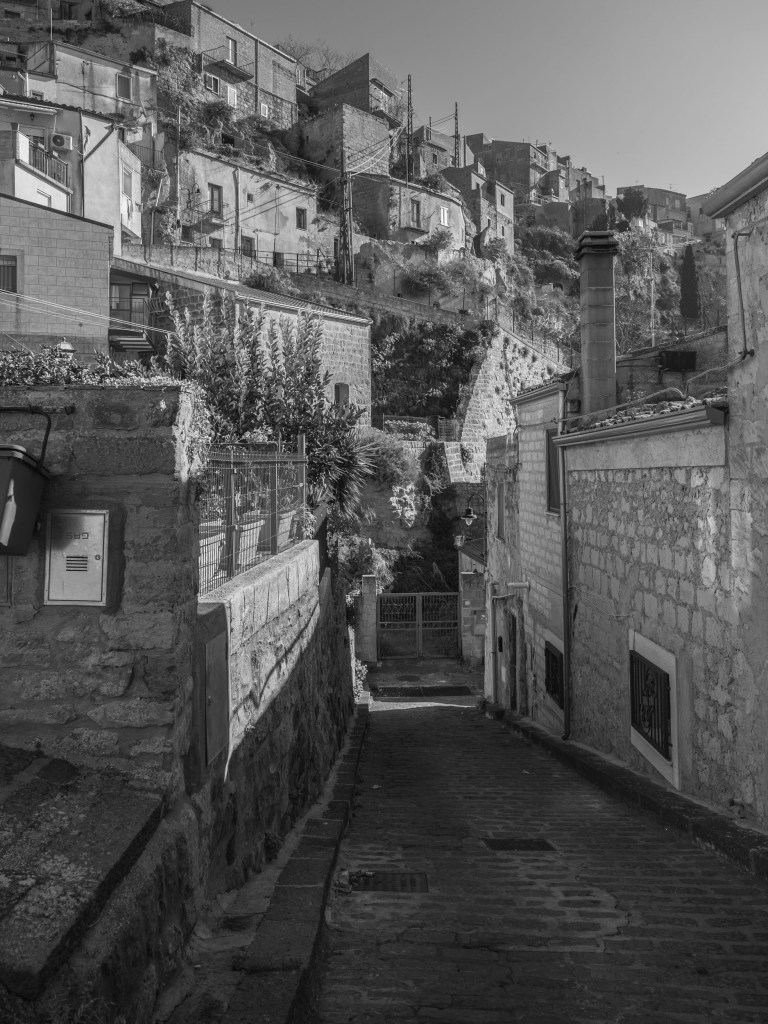 A narrow cobbled street descends between stone buildings, while houses rise in successive layers along the hillside beyond.