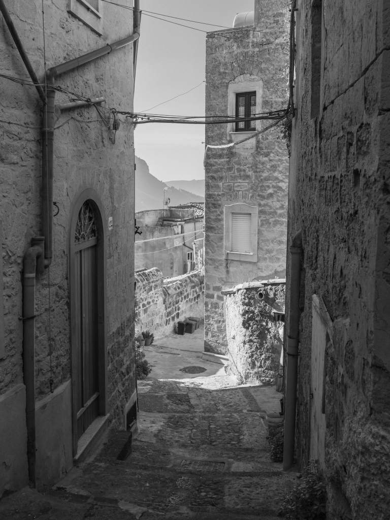 A steep passage between weathered stone houses opens towards distant mountains framed by walls, windows and overhead cables.