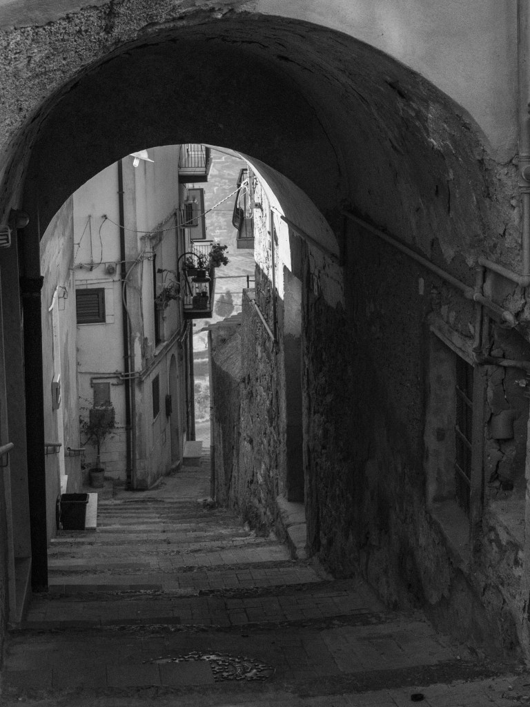 A dark arched passage leads down through a narrow residential street towards a brighter opening in the distance.