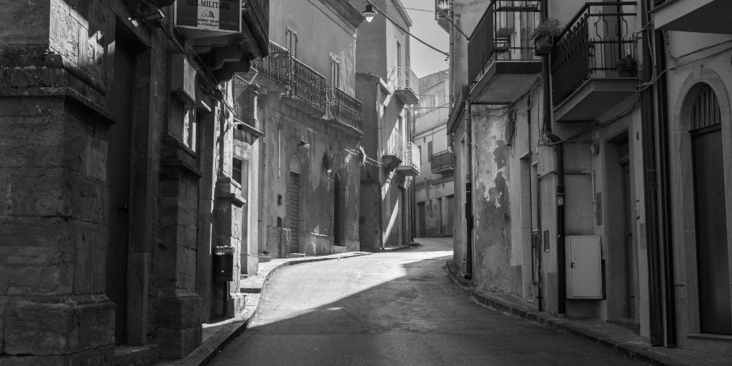An empty street curves uphill between worn façades and balconies, crossed by a sharp band of sunlight.