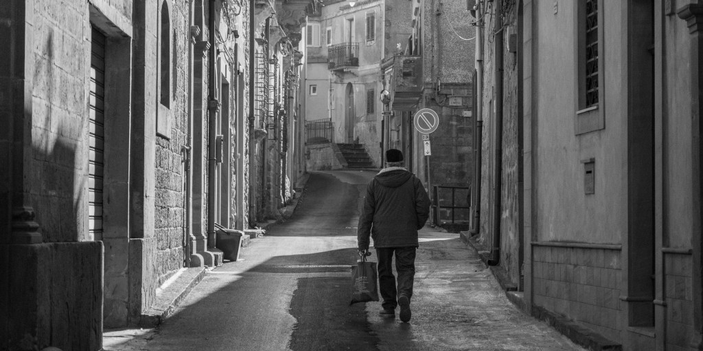 A solitary man carrying a shopping bag walks away along a quiet street between old buildings.