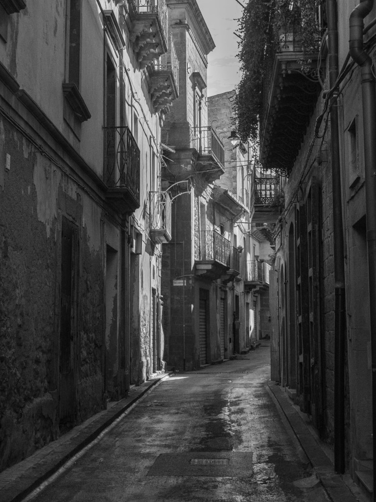 A shadowed street lined with balconies and worn façades curves towards a patch of light.