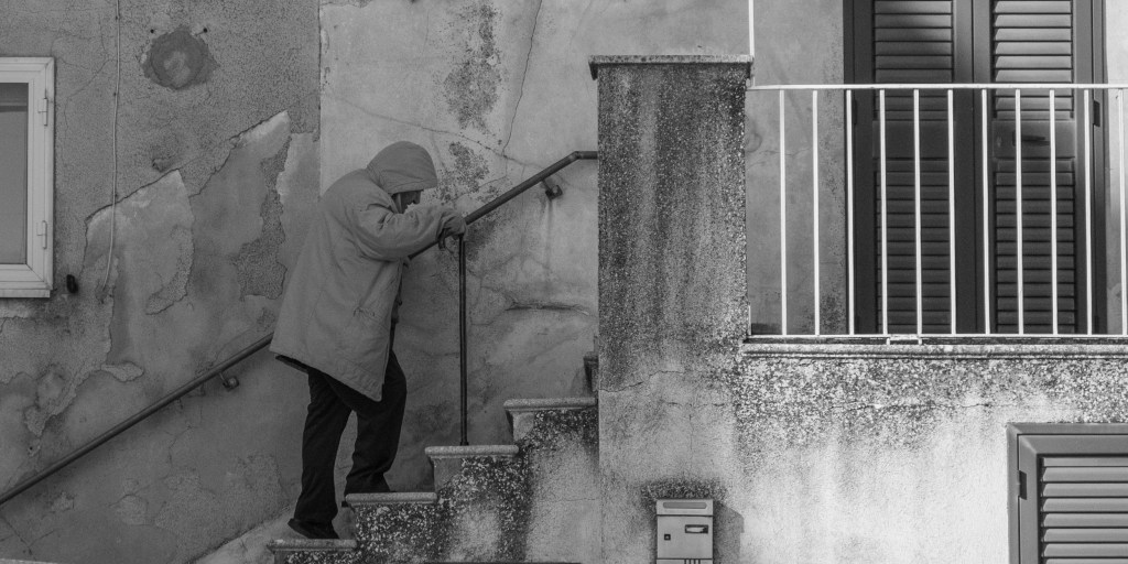 An older person using a walking stick climbs a short exterior staircase beside a weathered house.