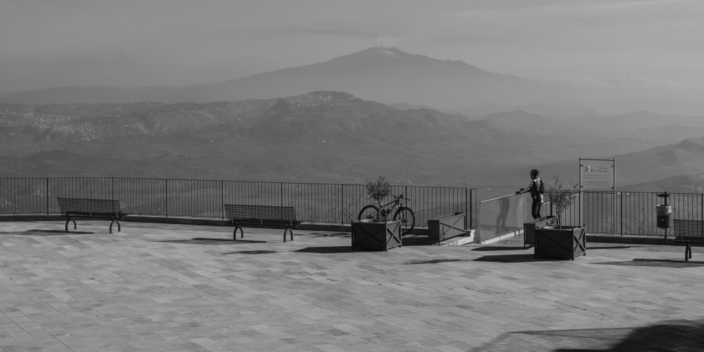 An open paved terrace overlooks distant mountain ranges, with benches, planters, a bicycle and a lone figure near the railing.