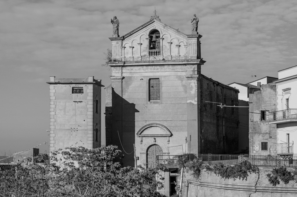 A weathered church façade rises above surrounding houses and vegetation beneath a lightly clouded sky.