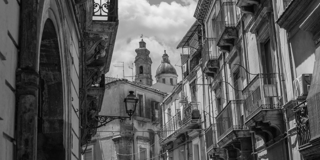 A narrow street in Caltagirone is lined with ornate balconies, while a church dome and bell tower rise above the rooftops.