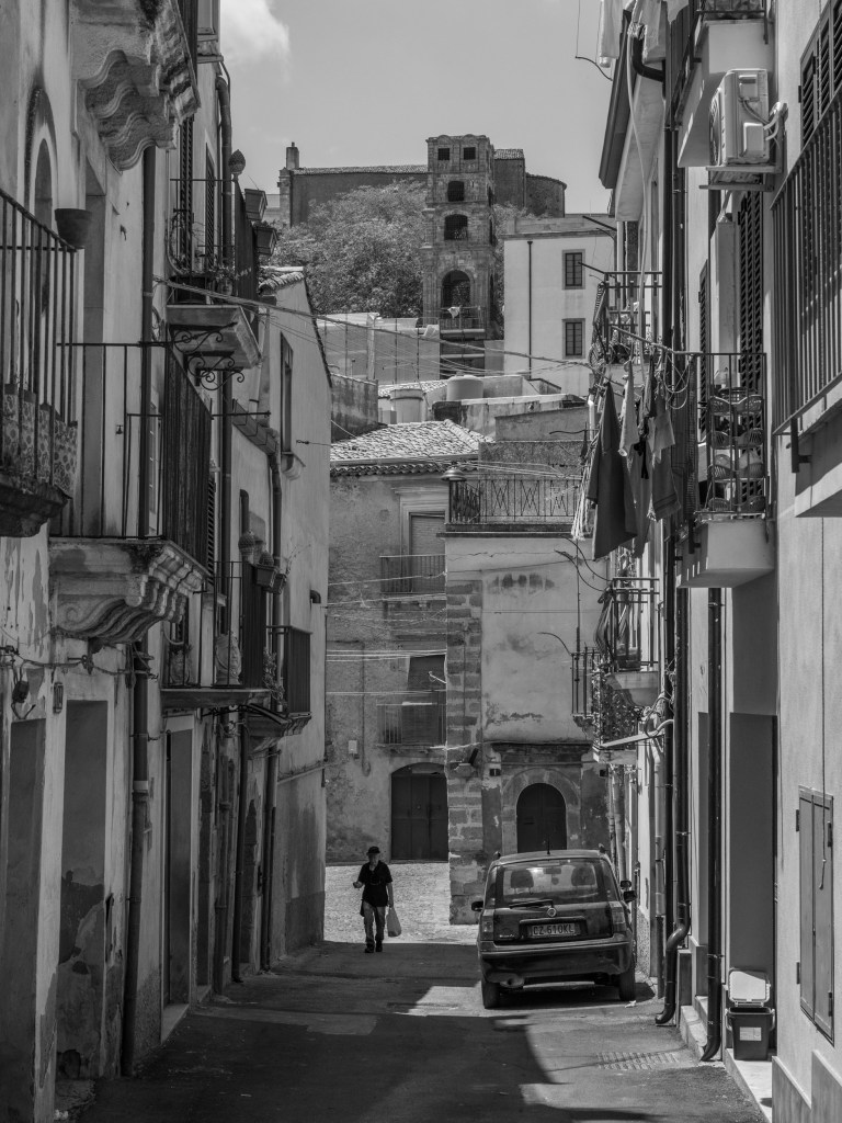 A pedestrian walks along a residential street with balconies, laundry and a parked car, beneath a large historic building in the distance.