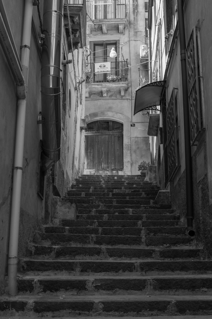 A steep stone staircase climbs through a narrow passage towards a large wooden door, framed by pipes, cables and balconies.