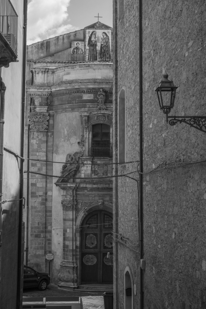An ornate church façade appears between two narrow walls, with religious mosaics, carved stonework and a traditional streetlamp.