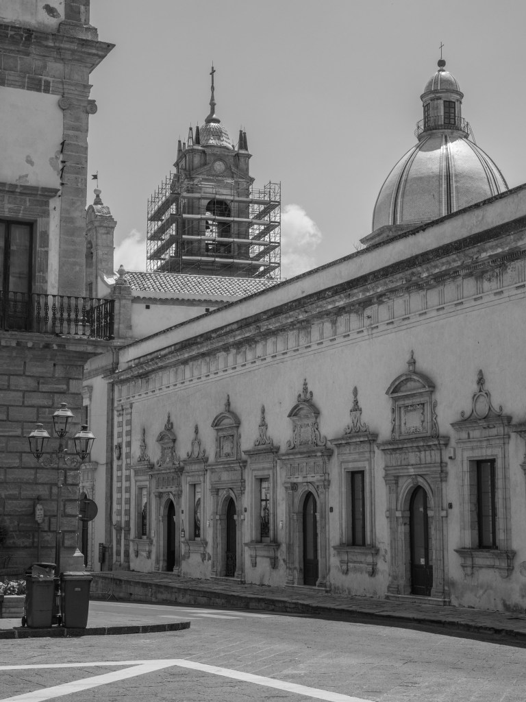 A long historic façade with repeated arched windows stands beneath a church dome and a bell tower covered in scaffolding.
