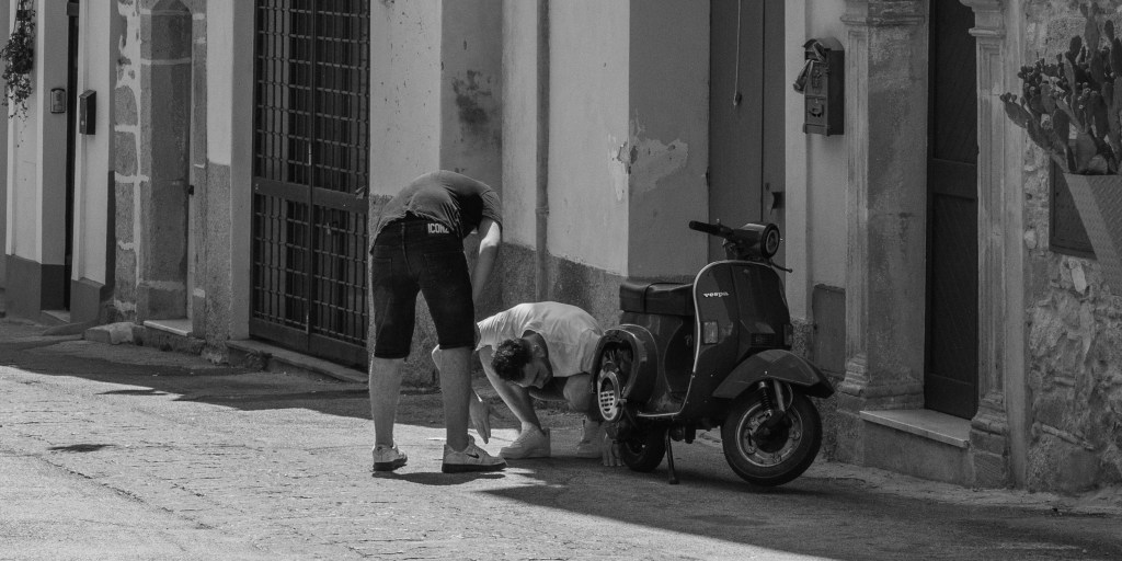 Two people bend beside a parked Vespa on a sunlit residential street.