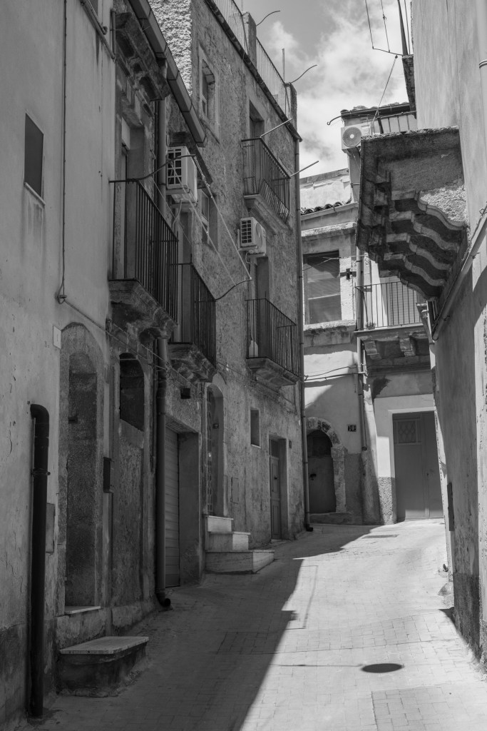 A quiet lane curves uphill between weathered buildings, balconies and air-conditioning units, divided by strong sunlight and shadow.
