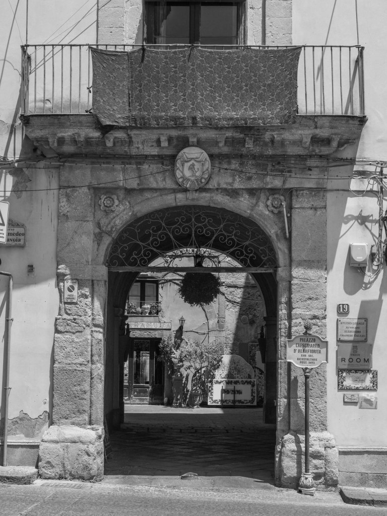 A large stone arch with decorative ironwork opens into a courtyard, beneath a balcony draped with patterned fabric.
