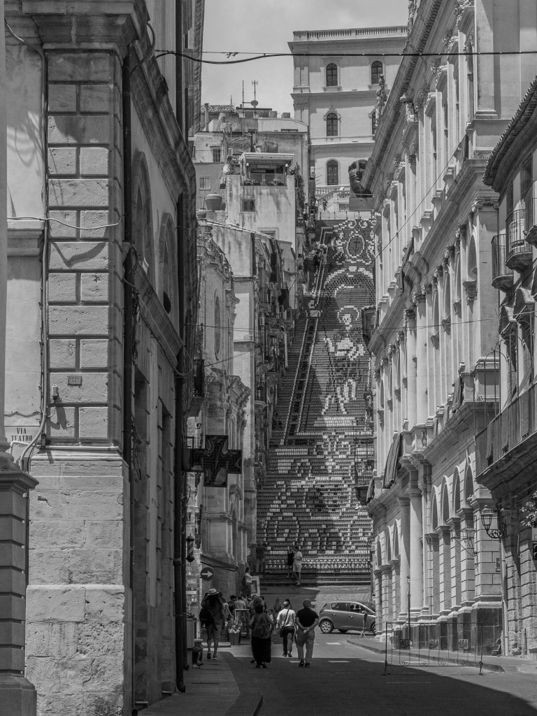 A long street leads towards Caltagirone’s monumental staircase, decorated with a large geometric pattern and framed by historic buildings.