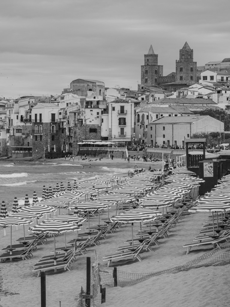 Rows of striped beach umbrellas fill the foreground, with Cefalù Cathedral rising above the town behind them.