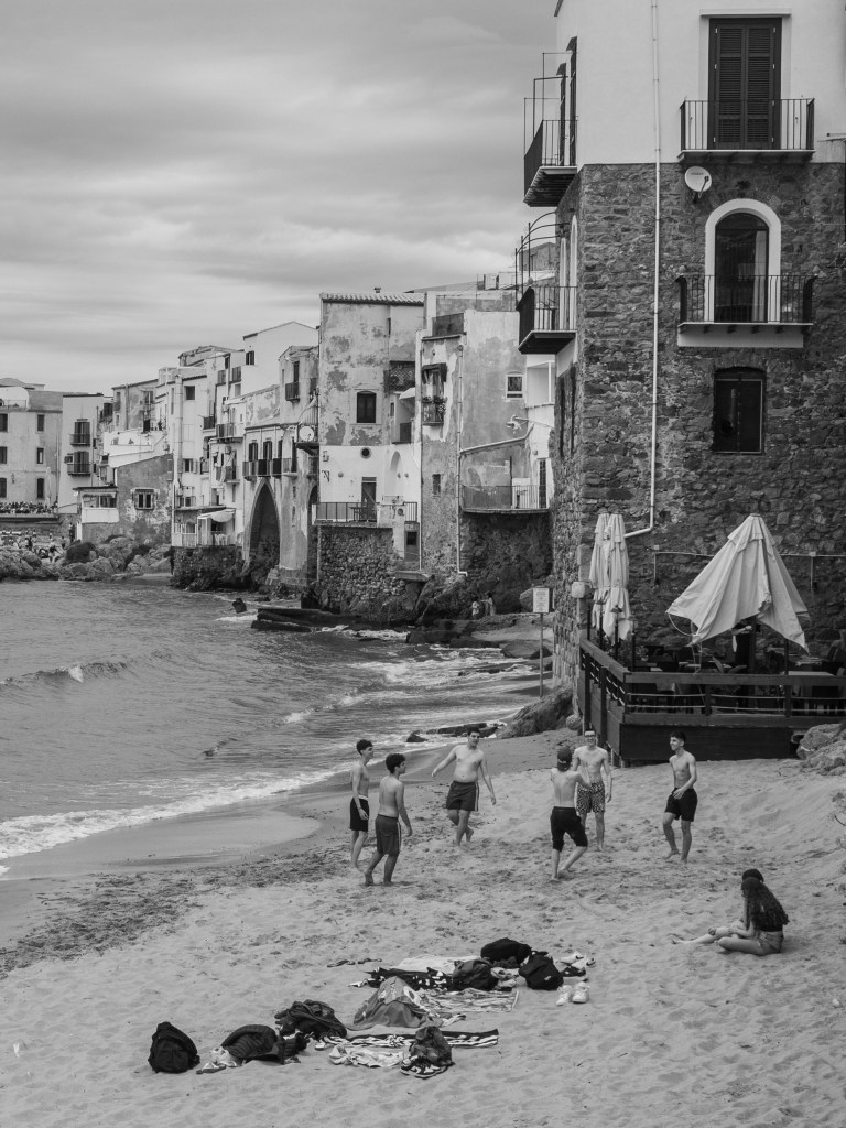 A group of young people plays on the sand beneath the houses of Cefalù’s historic waterfront.
