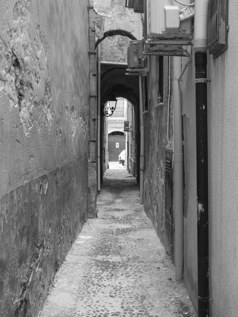 A narrow passage runs between weathered walls, arches, pipes and external air-conditioning units.