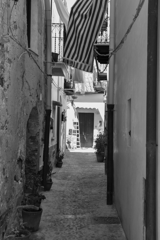 A striped awning hangs above a narrow residential alley with balconies, plants and a ladder.