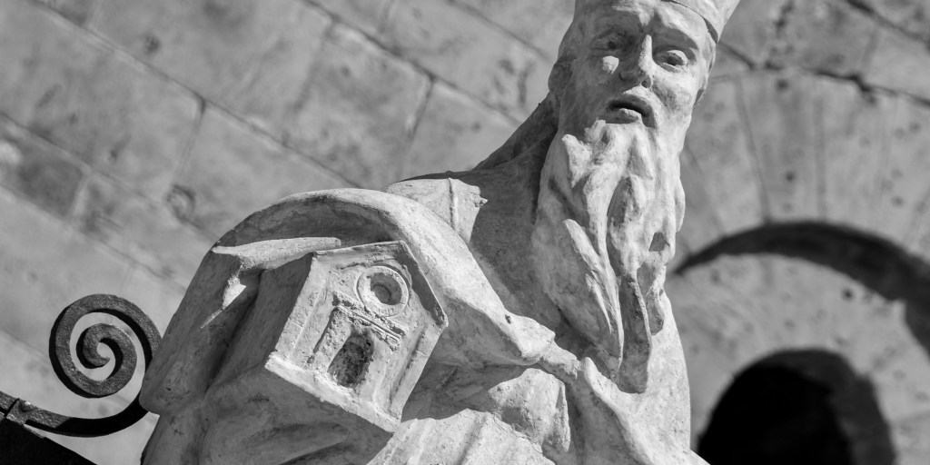 A stone statue of a bearded saint holds a small model of a church before the cathedral wall.