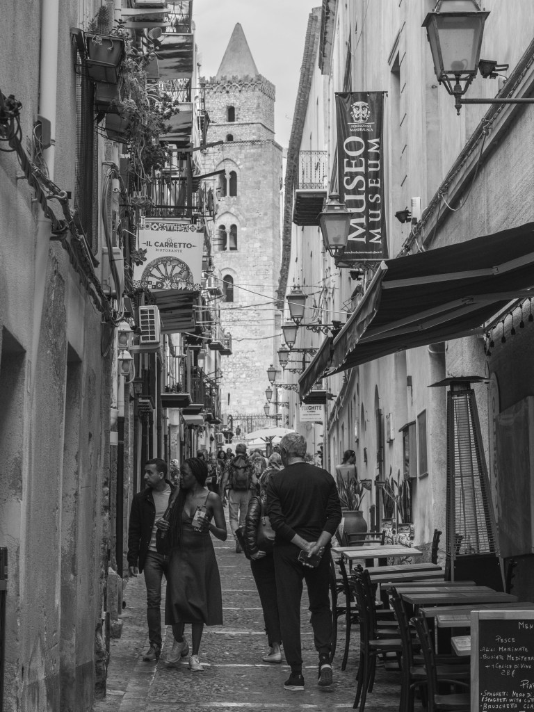 A crowded commercial street leads towards one of the towers of Cefalù Cathedral.
