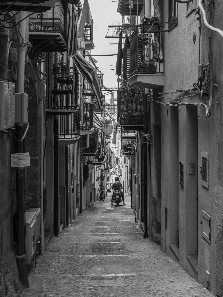 A scooter waits at the far end of a narrow lane enclosed by balconies, awnings and cables.