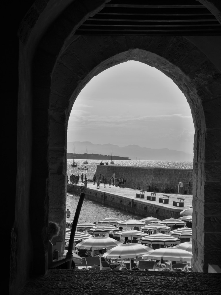 A large stone arch frames the harbour, crowds, boats and rows of beach umbrellas.