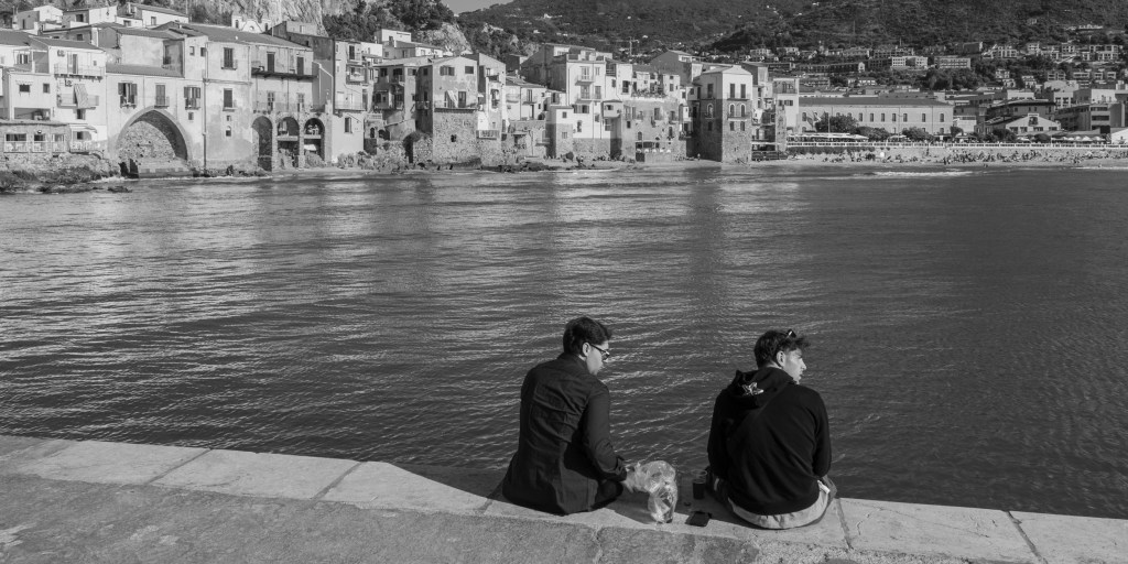 wo people sit on a stone seawall facing the water and Cefalù’s historic waterfront.