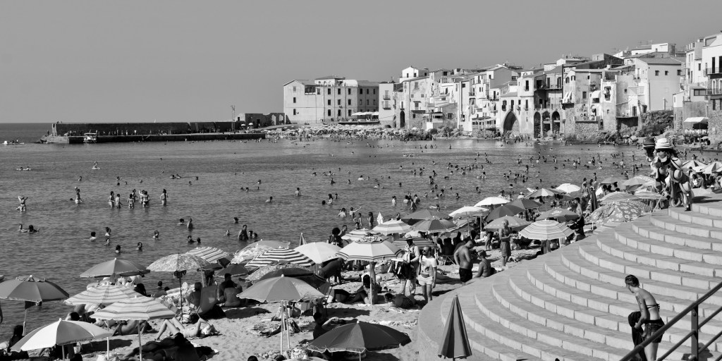 Crowds of swimmers and beachgoers fill the bay beneath the houses of Cefalù’s old town.