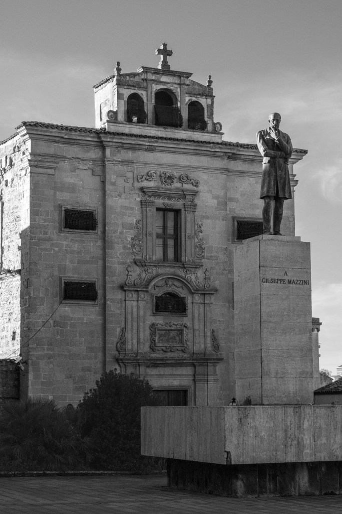 A statue of Giuseppe Mazzini stands before an ornate church façade and small bell structure.