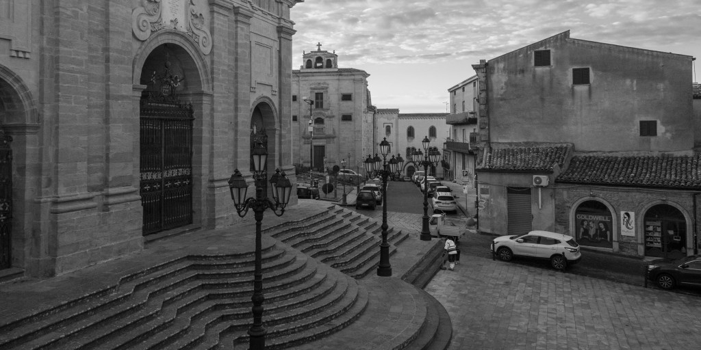 Curving cathedral steps descend into a square lined with decorative streetlamps, parked cars and historic buildings.