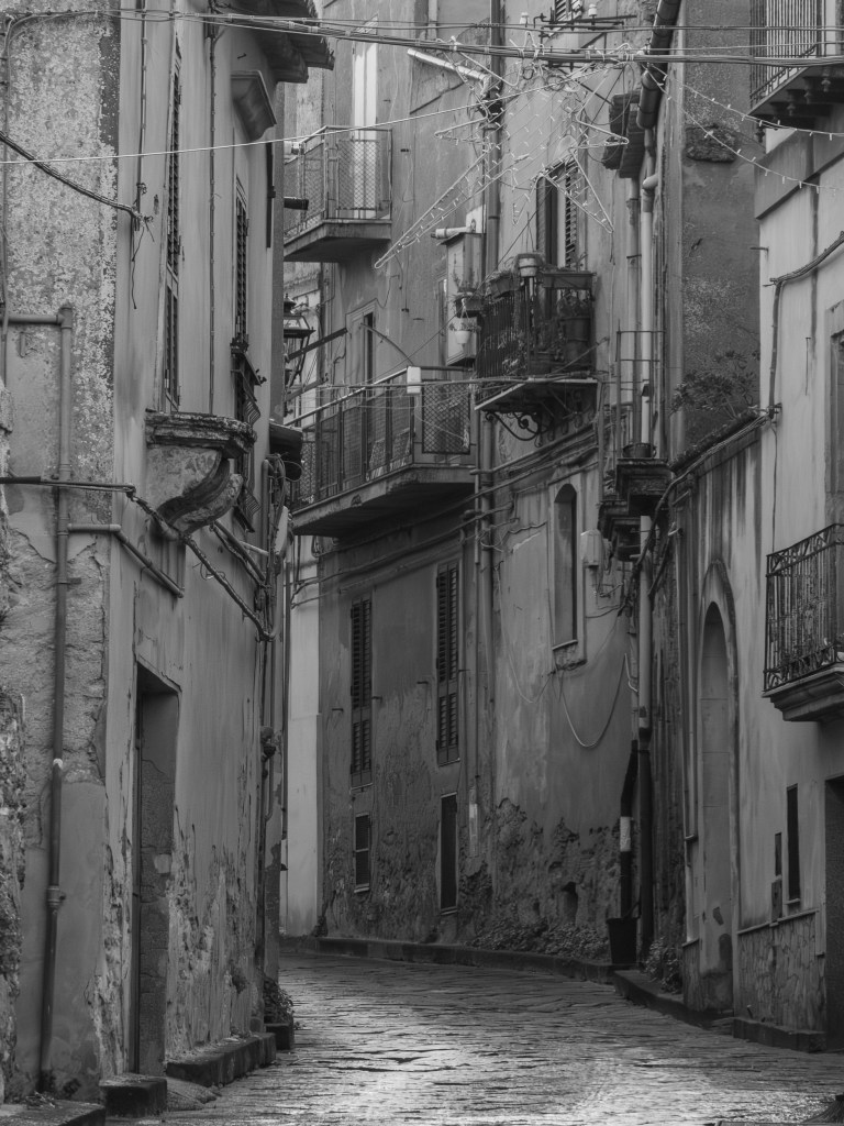 A narrow street rises between weathered façades, balconies and overhead cables beneath a suspended star decoration.