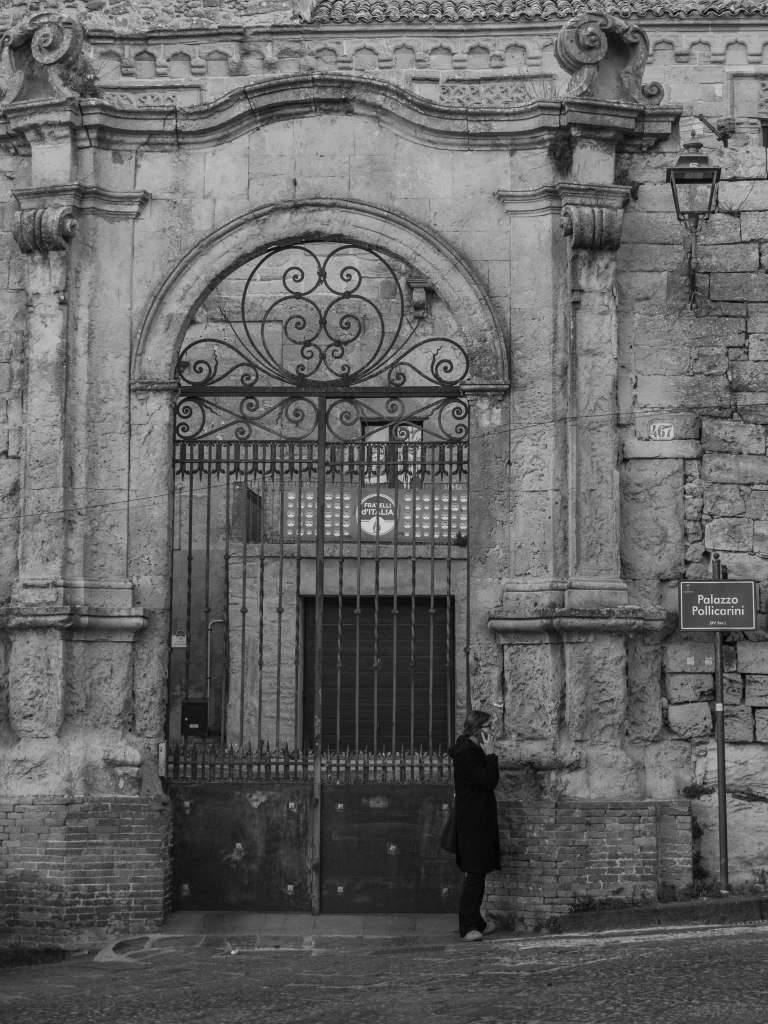 A solitary person stands beneath the monumental wrought-iron gate of Palazzo Pollicarini.