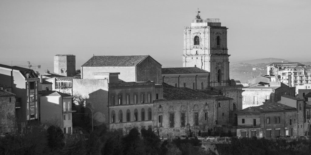 A cathedral and bell tower rise above surrounding buildings on the Enna skyline.