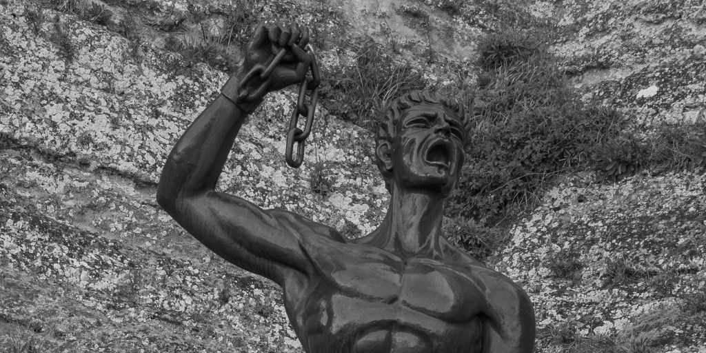 A bronze male figure raises a broken chain while shouting against a rocky background.