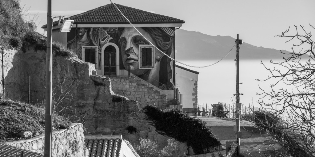 A building painted with a large human face stands above stone walls, rooftops and distant mountains.