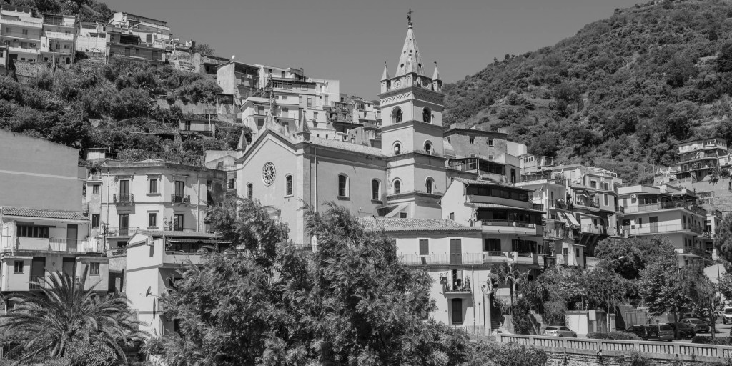 A hillside view of Itala, with a church and bell tower surrounded by densely layered houses and green slopes.